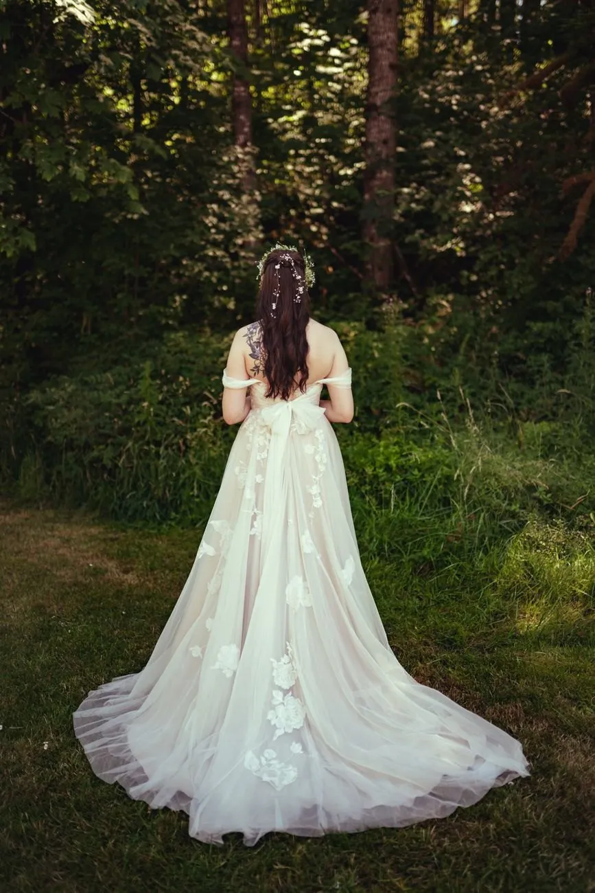 A bride seen from behind in a long dress, standing at the edge of a green wood