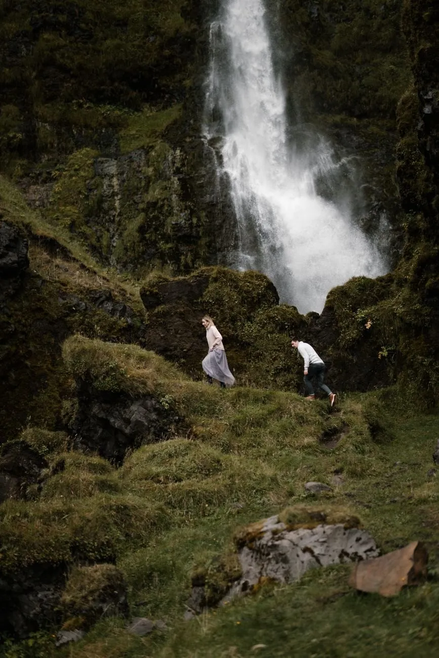 Two people climbing a mossy slope towards a tall waterfall