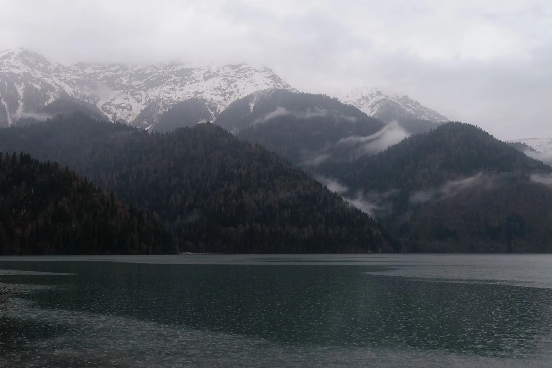 Dark wooded mountains with snow on the peaks, under low cloud above a still grey lake in western Norway