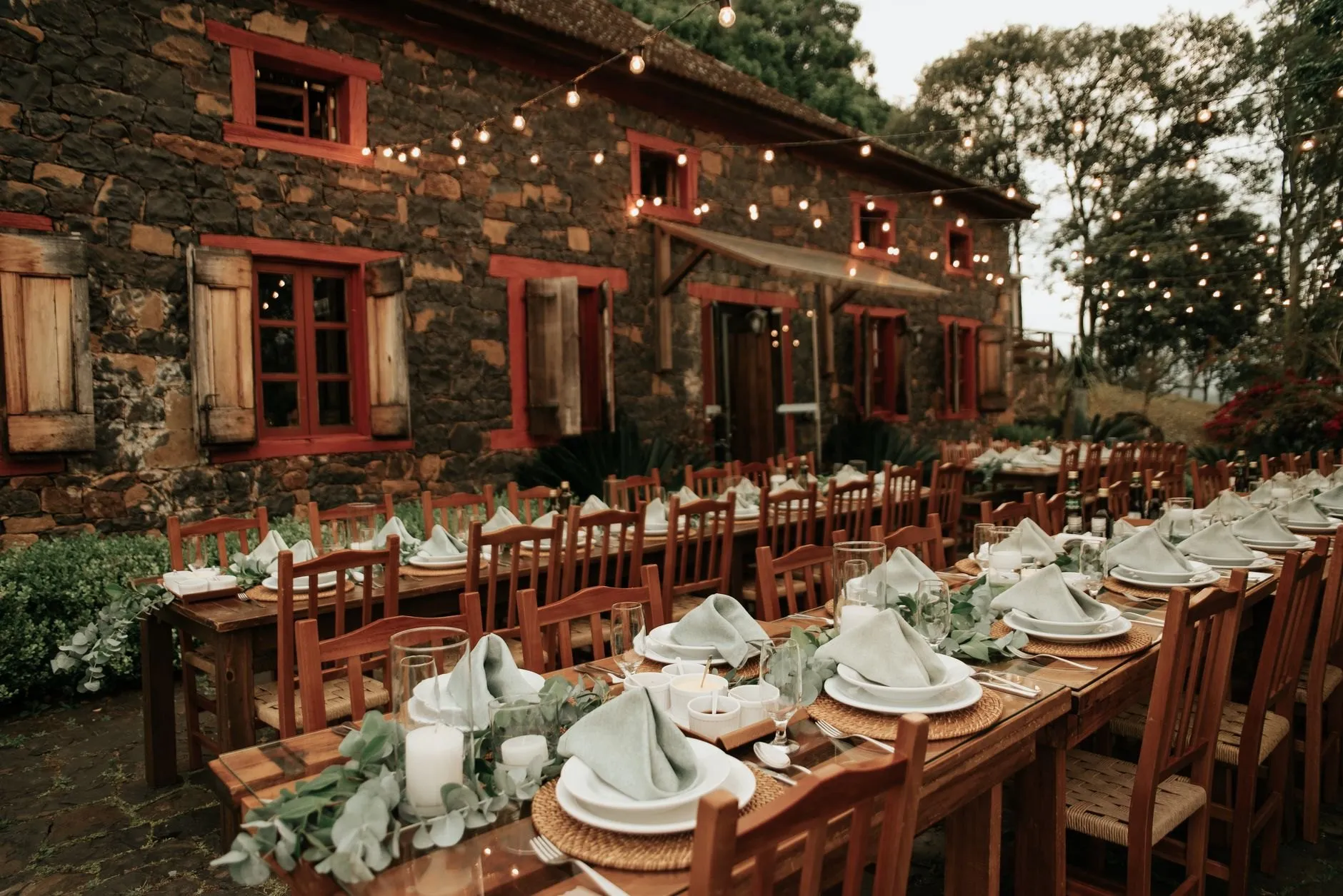 Long wooden tables set outside a stone farmhouse with red window frames under festoon lights